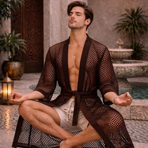 May include: A brown, open-weave robe with a tie waist. The robe is worn with off-white shorts. The image is set outdoors with a water feature and plants in the background. The robe is a stylish garment for lounging.