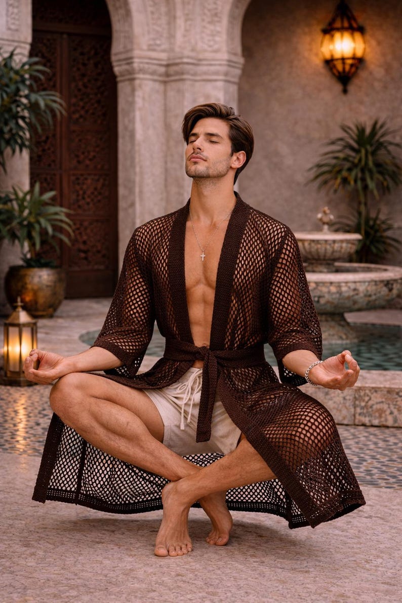May include: A brown, open-weave robe with a tie waist. The robe is worn with off-white shorts. The image is set outdoors with a water feature and plants in the background. The robe is a stylish garment for lounging.