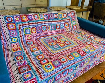 Op de afbeelding: Een kleurrijke, handgehaakte deken met een granny square design. De deken heeft een levendige mix van kleuren, waaronder roze, blauw, geel en rood. De deken is over een blauwe bank gedrapeerd, wat het ingewikkelde patroon en vakmanschap laat zien.