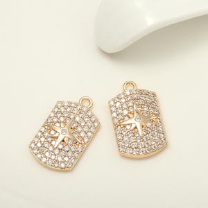 May include: Two gold-coloured rectangular pendants with a star design. The pendants are encrusted with small, clear crystals and feature a raised star in the centre. Each pendant has a small loop at the top for attaching to a chain or other jewellery component.