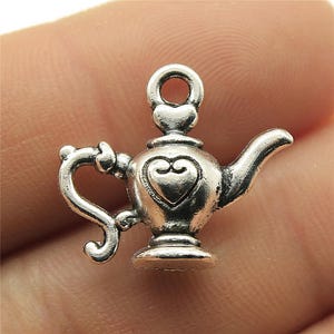 May include: A small, silver-colored charm in the shape of a genie lamp. The charm features a heart-shaped design on the front and a loop at the top for attaching to a necklace or bracelet. The handle and spout are curved.