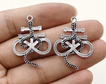 May include: Two silver-coloured pendants with a stylized design. Each pendant has a cross-like shape at the top, a snake winding through the centre, and an infinity symbol at the bottom. The pendants are held in a hand.