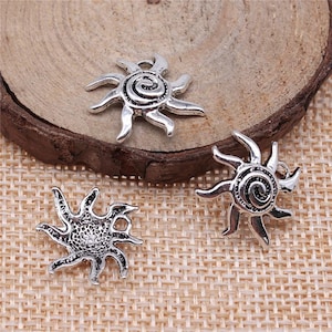 May include: Three silver-colored sun charms with spiral centers and pointed rays. The charms are displayed on a wooden slice and a woven beige surface, showcasing their intricate details and design.