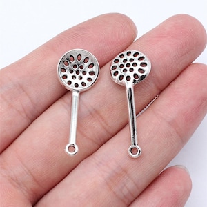 May include: Two silver-colored miniature slotted spoons. Each spoon has a round, perforated head and a long handle with a small hole at the end. These charms are likely for jewelry making or other crafts.