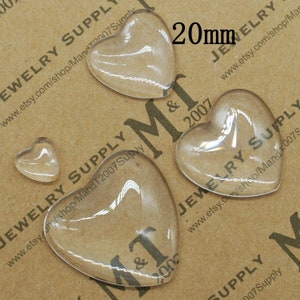 May include: Four clear glass heart-shaped cabochons, two are 20mm in size and two are smaller. The cabochons are on a brown surface.