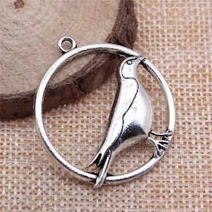 May include: A silver-colored bird charm pendant. The bird is perched inside a circular frame with a loop at the top for attaching to a chain or other jewelry. The charm has a detailed, antique finish.