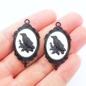 May include: Two black oval pendants with a raven design. Each pendant features a black raven perched on a branch against a white background, framed by a decorative black border. The pendants have a loop at the top for hanging.
