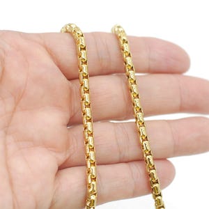 May include: A gold-toned box chain necklace featuring a series of interconnected, smooth, cylindrical links. The chain has a polished, reflective surface and a uniform, repeating pattern throughout its entire length.