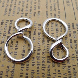 May include: Two silver-coloured infinity-shaped charms. Each charm features a figure-eight design, with a loop at the top and bottom connected by a curved section. The charms are displayed against a background of aged paper.