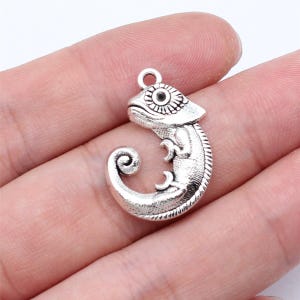 May include: A silver chameleon charm with intricate detailing. The charm has a curled tail and a loop for attaching to jewellery. The chameleon's eye is a prominent feature. The charm is made of a silver-colored metal.
