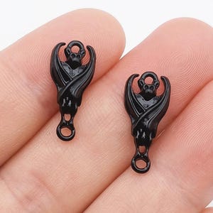 May include: Two black bat-shaped charms. Each charm features a bat with outstretched wings, a small loop at the top, and a larger loop at the bottom. The charms are made of a shiny, dark material.