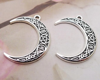 May include: Two silver-coloured crescent moon pendants. Each pendant features an intricate design with a pentagram and geometric patterns. The pendants have a loop at the top for hanging and are set against a soft, blurred background.