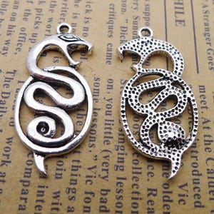May include: Two silver-coloured snake pendants. Each pendant features a stylised snake design within an oval frame. The snakes are coiled, with detailed heads and tails. The pendants have a loop at the top for hanging.