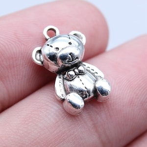 May include: A silver-colored teddy bear charm with a bow tie. The bear has a loop at the top for attaching to a necklace or bracelet. The charm is detailed with visible stitching and rounded features.