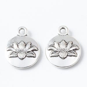 May include: Two silver-toned metal charms. Each charm is round with a raised lotus flower design in the center. A small loop is at the top for attaching to jewelry or other crafts. The charms have a vintage or antique appearance.