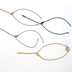 May include: Four delicate chain bracelets in rose gold, black, silver, and gold. The gold bracelet features a small, round, clear gemstone cluster. Each bracelet has an adjustable chain.