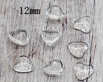 May include: Eight clear glass heart-shaped beads, each measuring 12 mm in diameter. The beads are arranged on a light brown wooden surface.