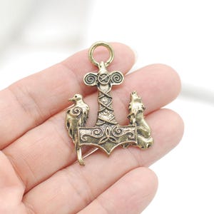 May include: A brass-toned metal pendant shaped like a Mjolnir hammer, featuring a central cross-hatched shaft, a top circular bail, and two flanking animal figures, a bird on the left and a wolf on the right, with intricate spiral engravings.
