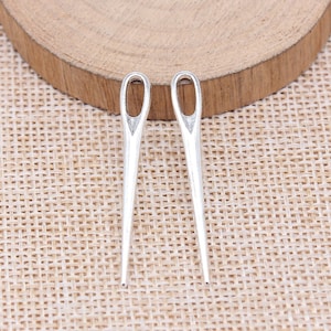 May include: Two silver-colored, large-eye sewing needles. The needles are positioned vertically, with their eyelets at the top. They are set against a textured, light brown background, suggesting a craft or sewing theme.