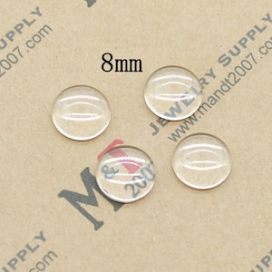 May include: Four clear glass cabochons, each measuring 8mm in diameter, are arranged on a brown paper surface. The cabochons are round and have a smooth, polished finish.