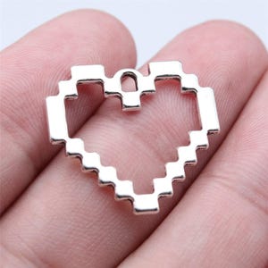 May include: A silver-colored, pixelated heart charm. The charm is made of metal and has a retro, digital design. The heart shape is open in the center and has a small loop at the top for attaching to a chain or other jewelry.