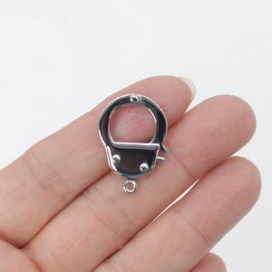 May include: A silver-toned metal jewelry clasp shaped like a miniature handcuff. The clasp features a circular opening, a hinged mechanism with two small protruding studs, and a small jump ring at the base for attaching to jewelry chains.
