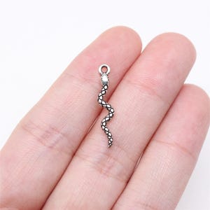 Peut inclure: Un charm en forme de serpent argenté avec un motif texturé en écailles. Le charm est en forme de serpentin incurvé, avec une petite boucle en haut pour l'attacher à un bijou. Le charm est tenu dans une main.
