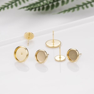 May include: Several gold-colored earring posts with flat, round settings are displayed. The posts have a simple design, with a small, circular base and a post for insertion. The settings are designed to hold decorative elements.