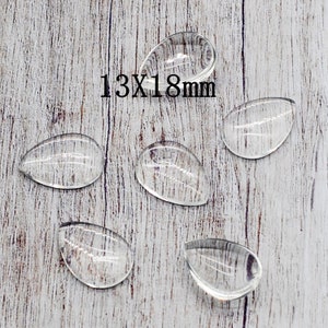 May include: Five clear glass teardrop-shaped cabochons, each measuring 13mm by 18mm, are arranged on a light wood surface.