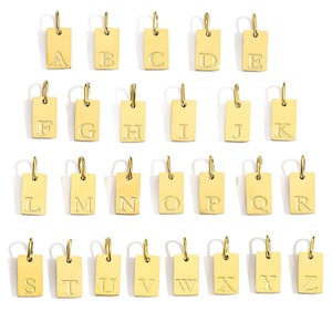 May include: A collection of gold-colored rectangular pendants, each engraved with a different letter of the alphabet from A to Z. The pendants have a small loop at the top for attaching to a chain or bracelet. The letters are in a simple, sans-serif font.