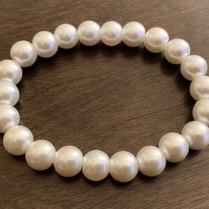 Puede incluir: Un brazalete de perlas blancas con un diseño sencillo. El brazalete está hecho de perlas redondas que están enhebradas en un cordón elástico transparente.
