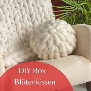 May include: A white knitted throw pillow with a large, round, flower-shaped design. The pillow is sitting on a white armchair. The text "DIY Box Bl&uuml;tenkissen" is written in red on a white background.
