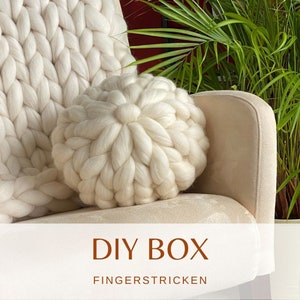 May include: A white knitted throw pillow with a large, round, puffy design. The pillow is sitting on a cream-colored armchair. The text "DIY BOX FINGERSTRICKEN" is displayed below the pillow.
