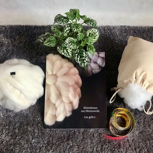 May include: A craft book titled "Bl&uuml;tenkissen aus Merinowolle Los geht's." with a white, pink, and gray flower-shaped wool cushion on the cover. There is a white wool cushion, a white cotton bag filled with white stuffing, and a measuring tape on a gray surface.