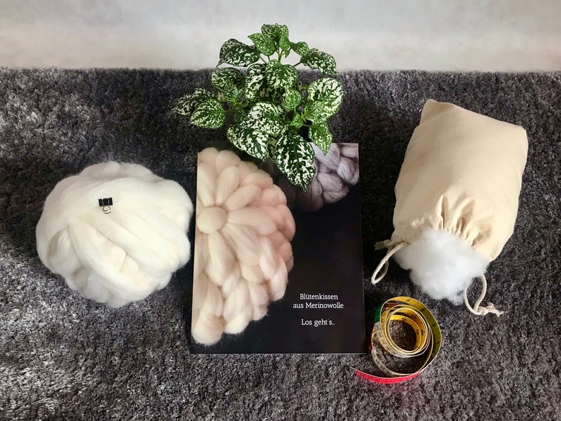 May include: A craft book titled "Bl&uuml;tenkissen aus Merinowolle Los geht's." with a white, pink, and gray flower-shaped wool cushion on the cover. There is a white wool cushion, a white cotton bag filled with white stuffing, and a measuring tape on a gray surface.