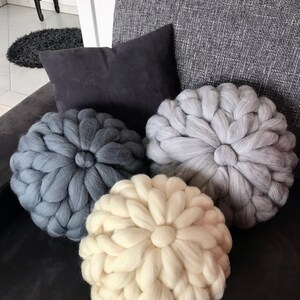 May include: Three round knitted throw pillows in a gray couch. The pillows are made of chunky yarn and have a flower-like design. The pillows are in shades of gray and white.