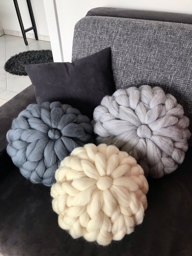 May include: Three round knitted throw pillows in a gray couch. The pillows are made of chunky yarn and have a flower-like design. The pillows are in shades of gray and white.