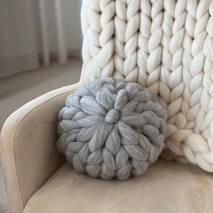 May include: A gray chunky knit throw pillow on a cream colored armchair with a white chunky knit throw blanket behind it.