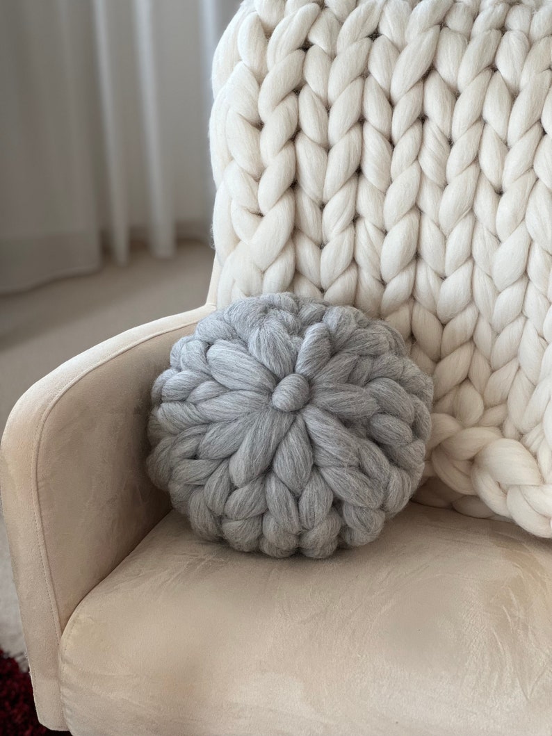 May include: A gray chunky knit throw pillow on a cream colored armchair with a white chunky knit throw blanket behind it.