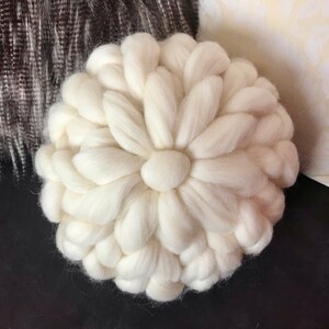 May include: A white, round, knitted pillow with a flower-like design. The pillow is made of thick yarn and has a soft, fluffy texture.