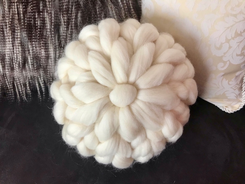 May include: A white, round, knitted pillow with a flower-like design. The pillow is made of thick yarn and has a soft, fluffy texture.