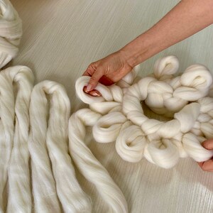 May include: A white yarn wreath being made by hand. The wreath is made of thick, fluffy yarn and is being tied into a circular shape. There are several strands of yarn lying on the table next to the wreath.