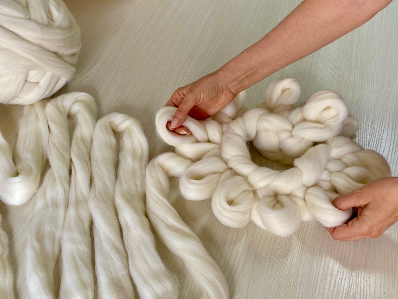 May include: A white yarn wreath being made by hand. The wreath is made of thick, fluffy yarn and is being tied into a circular shape. There are several strands of yarn lying on the table next to the wreath.