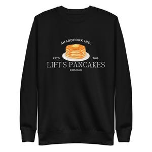 Può includere: Felpa nera con grafica di pancake con burro su un piatto. Il testo recita "SHARDFORK INC. ESTD 2016 LIFT'S PANCAKES ROSHAR". Un capo casual per gli amanti dei pancake.