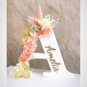 May include: A white wooden letter A decorated with pink and white dried flowers, gold butterflies, and the name Amelia written in cursive on the letter.