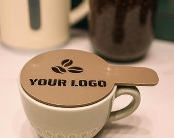 May include: A light green ceramic coffee cup topped with a flat, circular, tan-colored lid featuring a handle extension. The lid displays a dark brown graphic of three coffee beans and the text "YOUR LOGO" printed in a bold, sans-serif font.