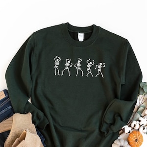 K&ouml;nnte beinhalten: Ein dunkelgr&uuml;ner Pullover mit einer wei&szlig;en Grafik von f&uuml;nf tanzenden Skeletten. Der Pullover liegt flach auf einem wei&szlig;en Hintergrund mit einem Paar braunen Stiefeletten, blauen Jeans und einem Zweig Eukalyptus mit Baumwollkn&auml;ueln.