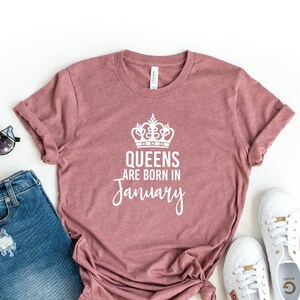 May include: Heather mauve t-shirt with a white crown and the text "Queens are born in January". The shirt is laid flat on a white background with a pair of denim shorts, sunglasses, and white sneakers.
