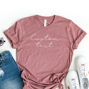 May include: A heather mauve t-shirt with the text "Custom text" in white script. The shirt is paired with distressed denim shorts, sunglasses, and white sneakers with gold accents.
