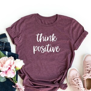 May include: Heather maroon t-shirt with the text "think positive" in white lettering. The shirt is shown with blue jeans, a bouquet of pink and white flowers, and pink trainers.